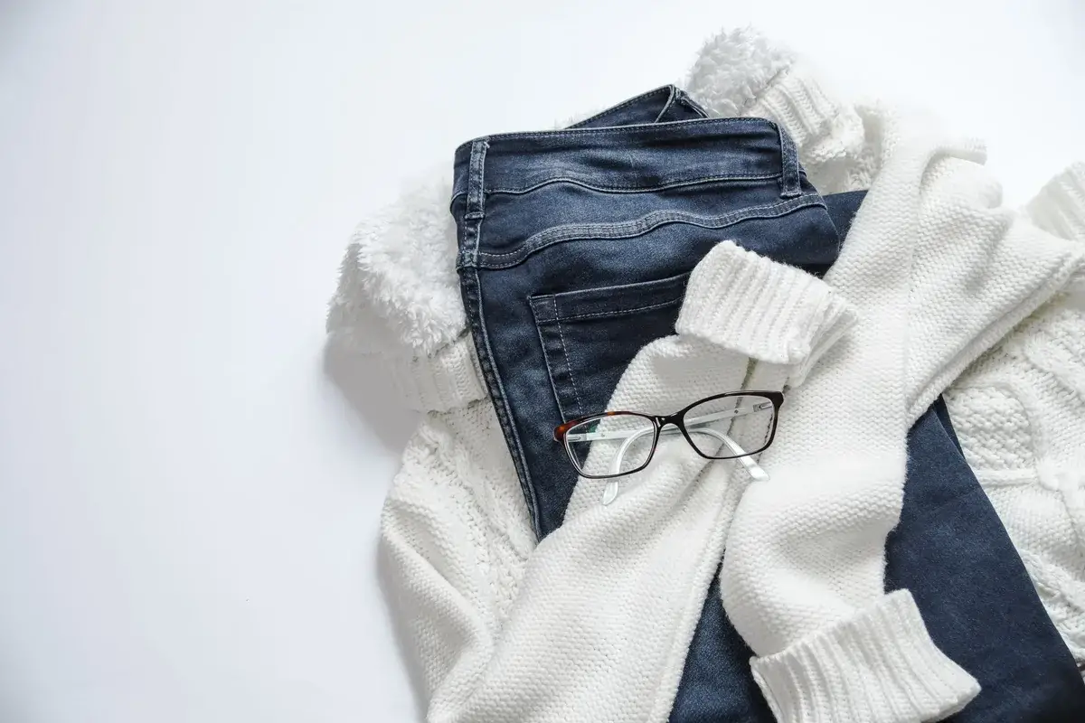 Pull en maille écrue, jean brut et lunettes posés à plat sur fond blanc